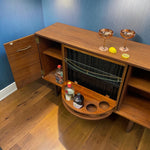 Load image into Gallery viewer, A wooden bar cabinet with an open door and a pull-out wine glass holder, styled on a wood floor against a blue textured wall with cocktail glasses and lemons on top.
