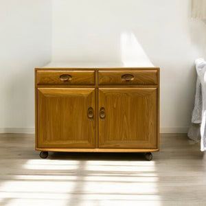 Wooden cabinet with two doors and a drawer on wheels in a room setting.