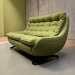 Load image into Gallery viewer, A mid-century modern low-profile loveseat sofa upholstered in textured olive green fabric, featuring a tufted backrest, curved arms, and a black metal swivel base, set against a concrete wall and light gray floor.
