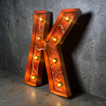 Load image into Gallery viewer, A large decorative letter K marquee light with glowing warm bulbs and an orange crackled glass face, leaning against a textured gray concrete wall.
