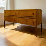 Load image into Gallery viewer, Mid-century modern wooden dresser with six drawers and tapered legs, positioned on a light wood floor in a room with warm sunlight and a window in the background.
