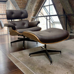 Load image into Gallery viewer, A mid-century modern lounge chair with matching ottoman in dark brown leather, set on a beige rug in a loft-style room with exposed brick walls and wood beams.
