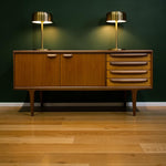 Load image into Gallery viewer, Mid-century modern wooden sideboard cabinet with two cabinet doors and three drawers, styled with two brass table lamps against a dark green wall and light wood floor.

