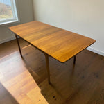 Load image into Gallery viewer, A mid-century modern wooden dining table with a warm honey-amber finish, shown in a room with hardwood floors and a window.
