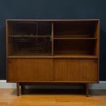 Load image into Gallery viewer, A mid-century modern wooden credenza or storage cabinet with a warm medium-brown wood finish, featuring two glass-front upper cabinets and two lower sliding doors, set against a dark blue wall and light wood floor.
