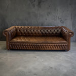 Load image into Gallery viewer, A brown leather Chesterfield sofa with deep button tufting, rolled arms, and a padded seat cushion, photographed against a textured gray concrete wall and floor.
