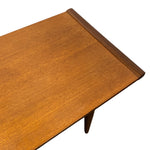 Load image into Gallery viewer, Close-up of a mid-century modern wooden table corner showing a warm medium-brown wood grain tabletop with a darker wood trim edge and a visible tapered leg against a white background.
