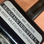 Load image into Gallery viewer, Close-up of a black metal hardware component with a white label reading 'SKOGHAUG INDUSTRI AS' and patent text, set against a warm brown leather background.
