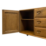 Load image into Gallery viewer, A mid-century modern wooden sideboard cabinet with a warm oak finish, featuring an open door, a single interior shelf, three drawers with oval pulls, and small caster wheels at the base, shown against a plain white background.
