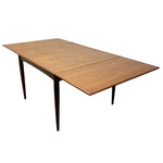 Load image into Gallery viewer, A mid-century modern extendable wooden dining table with a warm medium-brown wood grain top and dark tapered legs, shown on a plain white background.
