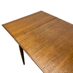 Load image into Gallery viewer, A close-up of a wooden dining table with a drop-leaf design, showing the warm medium-brown wood grain and a single leg against a plain white background.
