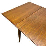 Load image into Gallery viewer, Close-up of a wooden dining table with a warm medium-brown finish and visible wood grain, showing part of the tabletop and a leg against a plain white background.
