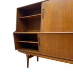 Load image into Gallery viewer, A mid-century modern wooden credenza or sideboard cabinet with warm medium-brown wood grain, featuring two sliding doors with slim dark handles, open shelves, and tapered legs, shown against a plain white background.
