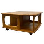 Load image into Gallery viewer, A mid-century modern wooden coffee table with a warm honey finish, featuring open side compartments and dark caster wheels, shown against a plain white background.
