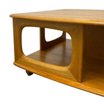 Load image into Gallery viewer, A close-up of a mid-century modern wooden coffee table on a white background, showing its warm honey-toned wood grain, curved edges, and open lower shelf.
