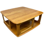 Load image into Gallery viewer, A mid-century modern wooden coffee table with a warm honey-amber finish, featuring a rounded square top, open lower shelf, and sculptural curved legs, shown on a plain white background.
