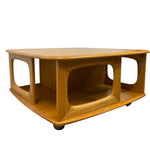 Load image into Gallery viewer, A mid-century modern wooden coffee table with a warm honey-amber finish, featuring curved sculptural legs, an open lower shelf, and small black casters at the base, shown on a plain white background.
