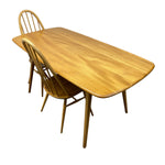 Load image into Gallery viewer, A mid-century modern wooden dining table with a warm honey finish, accompanied by two matching spindle-back chairs, isolated on a white background.
