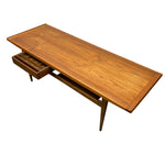 Load image into Gallery viewer, Mid-century modern wooden writing desk with a warm medium-brown wood grain finish, featuring a single drawer with a geometric patterned front and a lower open shelf, shown on a plain white background.
