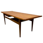 Load image into Gallery viewer, A mid-century modern wooden writing desk with a warm medium-brown finish, featuring tapered legs, a lower open shelf, and a subtle shadow box detail under the desktop, shown against a plain white background.

