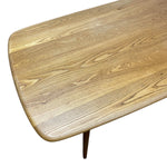 Load image into Gallery viewer, Close-up of a mid-century modern wooden table with a rounded rectangular top, displaying a warm honey-toned wood grain finish and a dark tapered leg against a white background.
