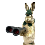 Load image into Gallery viewer, A rabbit-shaped novelty item, likely a glove, sock, or puppet, featuring a beige and brown fur pattern, wearing a dark green hat and bow tie, holding a black double-barrel shotgun against a solid white background.
