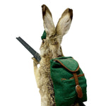 Load image into Gallery viewer, A taxidermy rabbit figurine shown from the back, wearing a green felt backpack with brown leather-like trim and a matching green hat, holding a black object against a plain white background.
