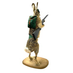 Load image into Gallery viewer, A taxidermy rabbit figurine standing on a wooden base, wearing a green backpack and holding a rifle, photographed against a plain white background.
