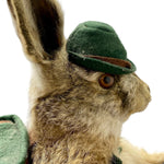 Load image into Gallery viewer, A close-up of a brown and beige rabbit wearing a small dark green felt hat with a brown band, shown against a plain white background.
