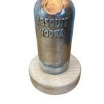 Load image into Gallery viewer, A vintage Absolut Vodka metal bottle stopper mounted on a round light wood base, shown against a plain white background.
