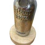 Load image into Gallery viewer, A vintage Absolut Vodka bottle-shaped decorative object with a distressed metallic finish, mounted on a round light wood base against a plain white background.
