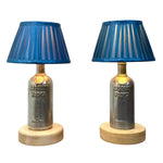 Load image into Gallery viewer, A pair of table lamps made from Absolut Vodka bottles, featuring blue pleated fabric shades, metallic silver-gray bottle bodies, and round light wood bases, set against a plain white background.
