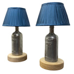 Load image into Gallery viewer, A pair of table lamps made from repurposed Absolut Vodka bottles, featuring dark gray metallic bodies, blue pleated fabric shades, and round light oak wooden bases, shown against a plain white background.
