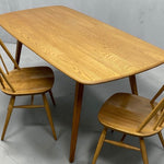 Load image into Gallery viewer, A light oak wood dining table with rounded corners and tapered legs, accompanied by two matching wooden chairs with spindle backs, shown on a concrete floor against a gray wall.
