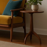 Load image into Gallery viewer, A mid-century modern wooden side table with a round top and three curved legs, styled next to a yellow armchair with a teal pillow and a potted green plant on top, set in a warm living room environment.
