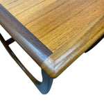 Load image into Gallery viewer, Close-up of a wooden table corner showing a warm medium-brown wood grain top with a darker brown beveled edge and a dark metal curved leg on a white background.
