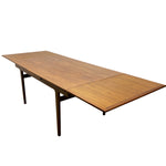 Load image into Gallery viewer, A mid-century modern extendable wooden dining table with a warm medium-brown wood grain top and dark metal legs, shown on a plain white background.
