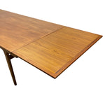 Load image into Gallery viewer, A close-up of a wooden extendable dining table showing the grain of the teak wood on the tabletop and a portion of the dark wood leg, isolated on a white background.
