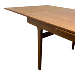 Load image into Gallery viewer, A close-up of a mid-century modern wooden dining table with a warm brown walnut finish, showing the extendable leaf panel seam and a drawer front on the side, isolated on a white background.
