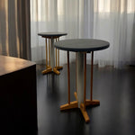 Load image into Gallery viewer, Two round tables with wooden legs on a dark floor with light filtering through curtains.
