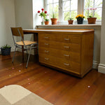 Load image into Gallery viewer, Wooden desk with drawers in a room with a window and potted plants.

