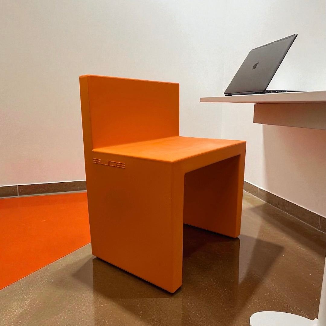 Orange chair with 'Slee' branding next to a desk with a laptop on a white wall background