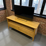 Load image into Gallery viewer, Wooden TV stand with a television on top against a brick wall with large windows.
