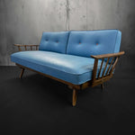 Load image into Gallery viewer, Blue sofa with wooden armrests against a gray concrete wall.
