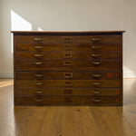 Load image into Gallery viewer, A vintage wooden multi-drawer storage chest with brass handles and label slots, set on a wooden floor against a plain wall with natural sunlight casting shadows.
