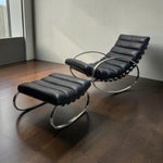 Load image into Gallery viewer, A modern black leather lounge chair with an attached matching ottoman, both featuring curved polished metal bases, photographed in a minimalist room with dark wood floors and a light gray wall.
