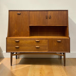 Load image into Gallery viewer, Mid-century modern wooden sideboard cabinet with a warm medium-brown wood grain finish, featuring a hutch upper section with two doors, an open central shelf, three drawers, and a lower cabinet door, set on tapered legs against a plain wall and light wood floor.
