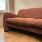 Load image into Gallery viewer, A close-up of a mid-century modern loveseat sofa upholstered in a textured rust-orange bouclé fabric, set on a light wood floor next to a window.

