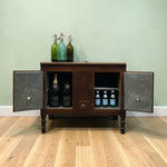 Load image into Gallery viewer, A dark wood sideboard cabinet with two open metal doors, styled with vintage soda bottles on top and wine and beer bottles inside, set against a sage green wall and light wood floor.
