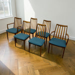 Load image into Gallery viewer, Six mid-century modern wooden dining chairs with teal upholstered seats arranged on a light oak herringbone floor against a white wall.
